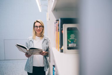 Barcelona 'da üniversite kütüphanesinde kitaplığın yanında durup kitap okurken gülümseyen ve kameraya bakan gözlüklü mutlu genç öğrenci