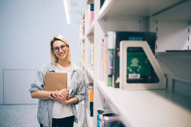 Kitapla gülümseyen ve kameraya bakan neşeli bayan öğrenci Barcelona 'daki çağdaş üniversite kütüphanesinde kitaplığa yaslanırken