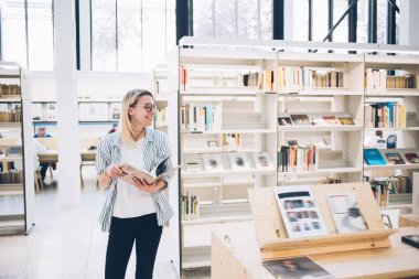 Zeki gündelik giyinen neşeli genç bir kadın sınav hazırlıkları için ders kitabında bilgi aramak için sırıtarak gülümsüyor. Halk kütüphanesindeki kitaplığın yanında gülen 20 'li yaşlarda mutlu bir hippi öğrenci.