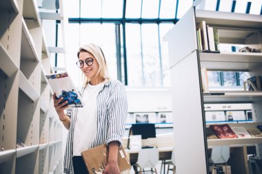 Mağazanın raflarının yanında ilginç açık kitaplarla duran mutlu beyaz kadın ve gülümseyen, eğitim edebiyatı okuyan zeki, sıradan bir kadın kütüphanede ödev için ders kitapları seçiyor.