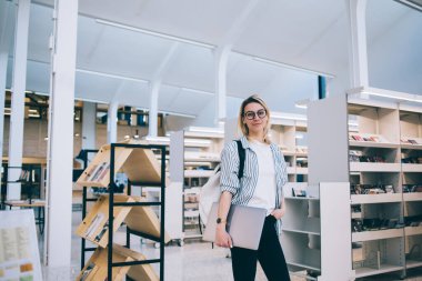 Kitabevinde dizüstü bilgisayarı olan başarılı beyaz kadın ressamın yarım boy portresi, 20 yaşında genç bir öğrenci çalışma alanındaki kitaplığın yanındaki kameraya bakıyor.