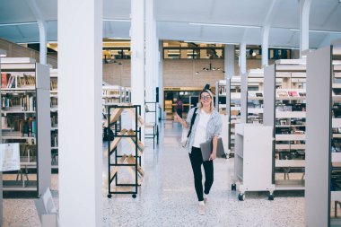 Optik gözlüklü mutlu bayan öğrencinin tam boy portresi kameraya gülümseyen ve başparmağını Ulusal Üniversite Kütüphanesinde gösteren dizüstü bilgisayarlı neşeli Kafkasyalı serbest yazar