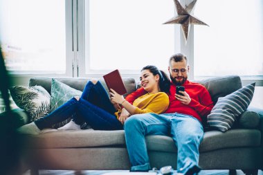 Gözlüklü ve günlük kıyafetleri olan mutlu bir adam akıllı telefon kullanıyor ve tatmin olmuş bir kadın rahat koltukta rahatlarken kitap okuyor ve açık modern oturma odasındaki büyük pencerelere karşı sevgiyle sarılıyor.