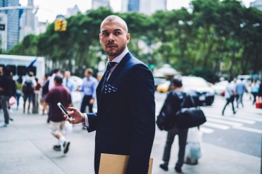 Lacivert takımlı, dosyasıyla ve akıllı telefonuyla kaldırımda yürüyen insanlarla bulanık arka planda duran çekingen işadamının yan görüntüsü