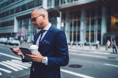 Elinde tablet ve kahve fincanı tutan kel bir iş adamı cep telefonuyla mesajlaşırken, yoğun bakışlı.