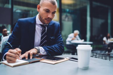 İş elbisesi ve kravatlı ciddi bir girişimci sokak kafesinde tabletle çalışıyor ve not defterine bir şeyler yazıyor. Masada içki içiyor.