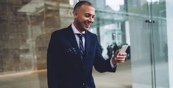 Happy male executive in navy blue suit standing with folder while text messaging on smartphone on blurred background with glass wall