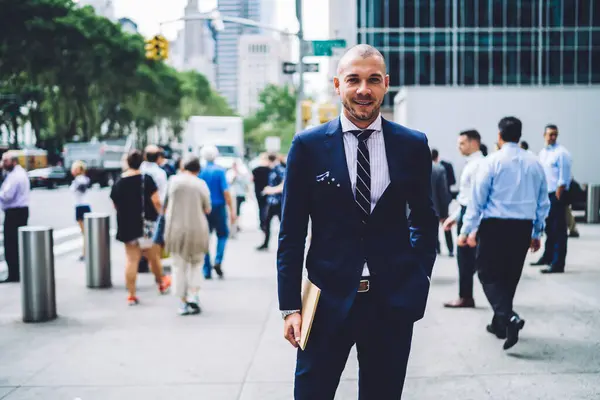 Portrait of confident successful professor 30 years old dressed in formal wear looking at camera, young Caucasian businessman standing at crowded metropolitan urban setting and posing in downtown