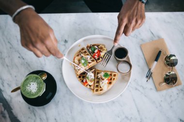 Etnik adamın ellerinin üstünde muzlu Belçika waffle 'ı ve kahvaltıda krema yerken yanında sağlıklı yeşil meyve püresi.