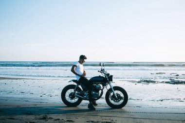 Güneş gözlüklü şık bir adam boş kumlu sahilde park etmiş motosikletli mavi gökyüzünde dalgalı bir sahil kenarında.