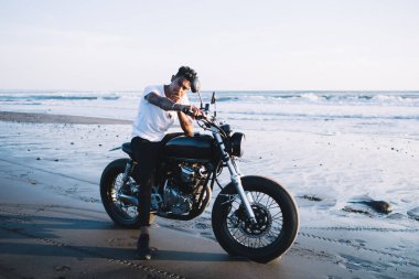 Güneş yanığı ve kıvırcık saçlı karizmatik motosikletçi gün batımında deniz sahilinde tek başına kameraya bakarken.