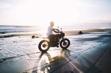Plaj boyunca motosiklet süren ve deniz manzaralı açık havada gün batımında ıslak kumda lastik izleri bırakan yüzü olmayan bir adamın yan görüntüsü.