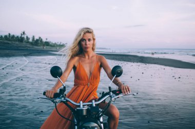 Turuncu elbiseli çekici genç bir bayan motosiklette oturuyor ve alacakaranlıkta deniz kıyısında kameraya bakıyor.