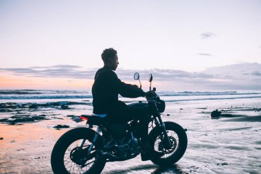 Dalgalı okyanus ve bulutlu gökyüzü boyunca ıslak sahilde motorsiklet süren güçlü İspanyol adamın yan görüntüsü