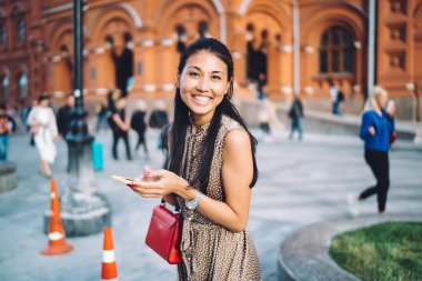 Elinde cep telefonu olan neşeli kadın turistin yarım boy portresi şehrin kenar mahallelerinde poz veren Asyalı turist modern akıllı telefonuyla kameraya gülüyor.