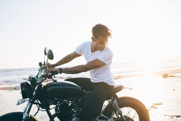 Hispanic handsome male biker in trendy glasses and light clothes driving motorbike along desert shiny beach near sea and looking down