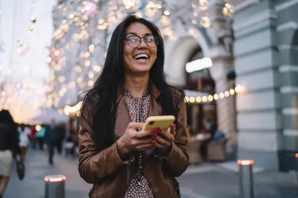 夜のウォーキング中に都市の設定で手で笑うセルラー技術を持つ陽気な女性ユーザー スマートフォンを保持し笑うビジョン補正のための古典的なアイウェアのヒップスターガール — ストック写真