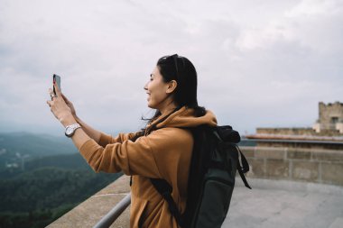 Gezide fotoğraf çekmek için cep telefonu kullanan neşeli Asyalı kadın blogcu, tatil turları sırasında konum videosu çeken olumlu milenyum kadın turist