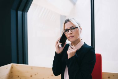 Resmi takım elbiseli ve gözlüklü profesyonel ciddi kadın yönetici telefonda konuşuyor ve modern çalışma alanının penceresinde otururken iş konularını tartışıyorlar.