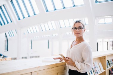 Barcelona 'daki modern kütüphanede akıllı telefon ve kitaptan veri ararken resmi kıyafet ve gözlüklü kendine güvenen bayan yönetici kameraya bakıyor.