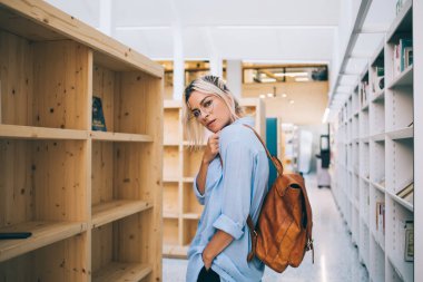 Barcelona 'da üniversite kütüphanesinde kitaplıklar arasında dururken sırt çantalı ve gözlüklü genç bir kadının kameraya baktığı yan bakış.