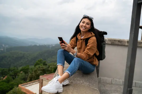 Portrait of smiling asian female traveler with backpack holding mobile phone chatting via roaming connection, happy woman tourist satisfied with roaming for share in blog media and multimedia content