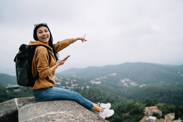 Happy asian female traveler with backpack excited with trip on vacations holidays using smartphone for blogging, positive woman explorer pointing on locations sitting on historical place setting