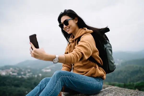 Happy hipster girl explorer in sunglasses shooting video for travel blog on mobile phone camera visiting historical place, smiling asian female tourist posing for selfie on smartphone during vacations