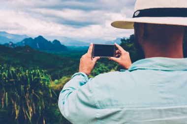 Erkek turistin cep telefonuyla doğa manzarasını çekerken ekran görüntüsüne odaklandığı arka plan görüntüsü. İnsanın, dağlara seyahat ederken manzaranın fotoğrafını çekerken, amaçsızca dolaşması.