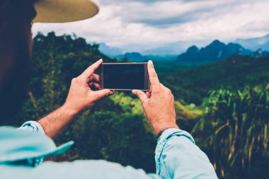 Erkek turistin cep telefonuyla doğa manzarasını çekerken ekran görüntüsüne odaklandığı arka plan görüntüsü. İnsanın, dağlara seyahat ederken manzaranın fotoğrafını çekerken, amaçsızca dolaşması.