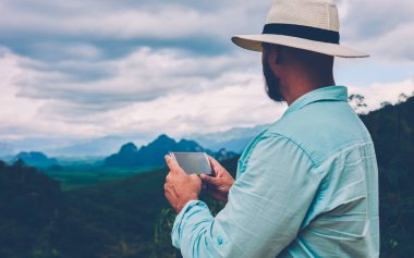 Erkek turist elinde akıllı bir telefonla ufuk çizgisine bakarken Tropikal Doğa 'yı ziyaret ediyor. Adam açık havada seyahat ederken sosyal ağ uygulaması üzerinden çevrimiçi video izliyor.