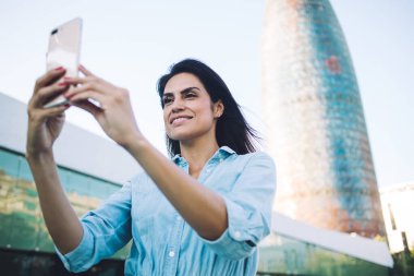 Şehir planında selfie çekmek için cep telefonu kullanan çekici beyaz kadın blogcu, şehirdeki sosyal ağlarda fotoğraf çekmek ve paylaşmak için akıllı telefon tutan oldukça olumlu bir kadın.