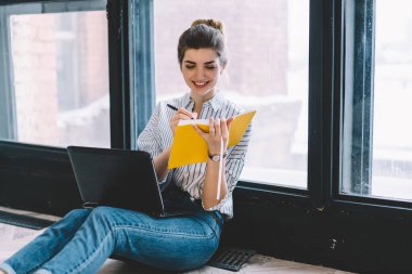 Öğrenmek için dizüstü bilgisayarda araştırma yaparken eğitim kitabında bilgi notları alan neşeli bayan öğrenci, netbook ile mutlu bir Kafkasyalı serbest yazar, not defteri kullanıyor.