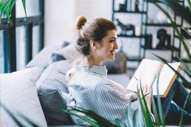 En iyi satan edebiyat kitapları satan neşeli kadın okuyucu. En sevdiği hobi için rahat koltukta dinleniyor. Yüzünde tatlı bir gülümseme, elinde eğitim kitabı olan beyaz bir kadın.