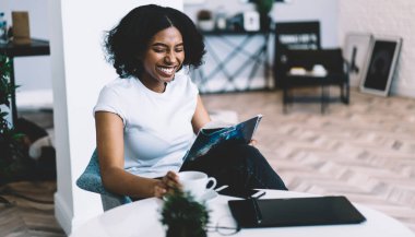 Neşeli Afrikalı Afrikalı Amerikalı ev hanımı kendi dairesinde oturmuş moda dergisinin yeni sayısını okuyor. Moda dergisi dergilerinde yeni basılmış derisi koyu tenli bir kadın. Fotokopi odası olan bir tişört giymiş.