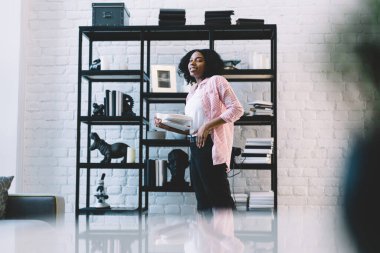 En çok satan edebiyatı elinde tutan modaya uygun giyinmiş kadın portresi ve hafta sonu modern apartman içinde poz veren, kara derili, kitabı kameraya gülümseyen kadın.