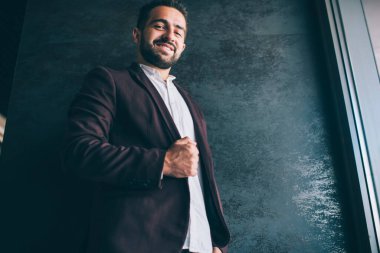 Neşeli, güler yüzlü, yakışıklı, etnik bir erkeğin şık takım elbisesinin altında çalışma alanında kameraya bakarak vakit geçirmesinden.