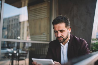 Ciddi sakallı, şık takım elbiseli ve modern binanın balkonunda otururken tablet kullanan kablosuz kulaklıklı bir etnik girişimci.