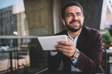 Kablosuz kulaklıklarla müzik dinlerken ve modern binanın balkonunda tablet kullanırken gülümseyen ve başka tarafa bakan takım elbiseli mutlu İspanyol adam.