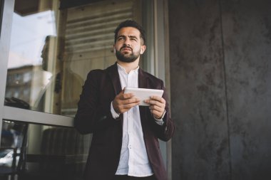 Tablet kullanan, müzik dinleyen, çağdaş binanın penceresinin yanında dikilen ve gözlerini kaçıran başarılı İspanyol takım elbiseli adam.