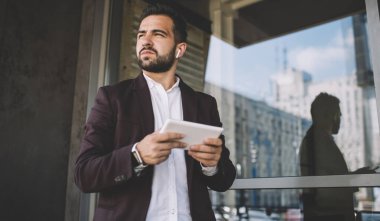 Resmi kıyafetli, düşünceli İspanyol bir erkek modern binanın penceresinin yanında durup tablet kullanarak kablosuz kulaklıkla müzik dinliyor.