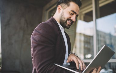 Modern laptopta gülümseyen ve göz gezdiren şık takım elbiseli sakallı etnik erkek güneşli bir günde şehrin dışında dikilip çalışırken...