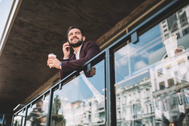 Mutlu gülümseyen, zarif takım elbiseli, temiz bir içecek içen, balkonda çitlere yaslanmış akıllı telefon kullanan, yüzünü çevirmiş, partneriyle konuşan etnik bir erkekten.