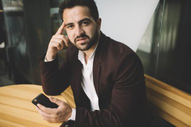 Kafası karışmış, kafası karışmış, akıllı telefonlu bir erkek çalışanın kahve dükkanında kameraya bakarken iş problemini kafa kafaya verip düşünmesi.