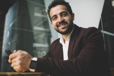 Kahve dükkanında kameraya bakarak bilek sörfü cep telefonunda akıllı saati olan pozitif sakallı başarılı bir menajerden.