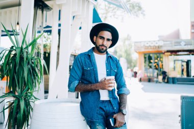 Gündüz vakti cafe terasında dikilirken sıcak içecek ve akıllı telefonuyla kameraya bakan modaya uyan kendine güvenen İspanyol serbest yazar.