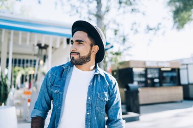 Modaya uygun, siyah şapkalı, mavi ve beyaz tişörtlü, güneşli bir günde gülümseyen ve sokakta hayal kuran son moda etnik erkek.