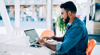 Etnik erkek uzaktan kumandalı işçinin modern açık hava kafeteryasında otururken boş ekran laptopuyla video izlerken yan görüntüsü