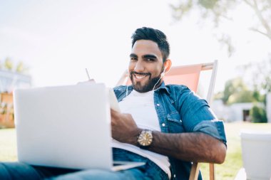 Akıllı içerikli rahat adam dizüstü bilgisayarlı ve kulaklıklı sandalyede güneşli şehir parkında kameraya bakıyor.