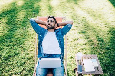 Zeki içerikli, sıradan kot gömlekli ve kot pantolonlu gülümseyen, parkta bir odada dizüstü bilgisayarda oturan etnik bir erkek. 